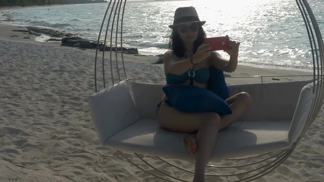 A young woman on a tropical beach sits on a swing, making a selfie.