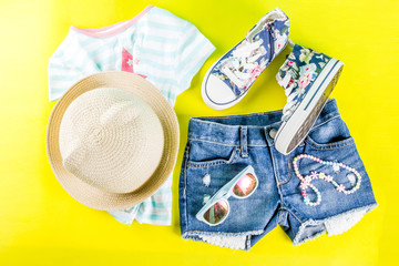 Summer vacation concept, summer child's cloth set - kids shorts, T-shirt, hat, sun glasses, bracelet necklace, sneakers, bright yellow background above flat lay copy space