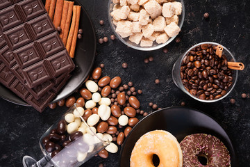 Glass with coffee beans next to chocolate tablets, donuts, brown sugar and other glass with peanuts in chocolate on dark wooden background