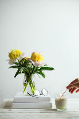 Cozy still life with peonies in a glass jar on a pile of books, glasses and woman's hand lighting the candle, selective focus, white background