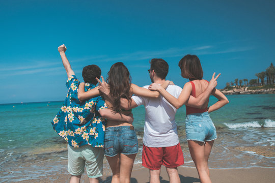 Cheerful young guys and girls are looking at the water with enjoyment. They are standing and embracing while turning back. Funny company entertainment at seaside concept 