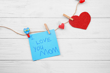 Card with words "I love you Mom", string and pegs on wooden background