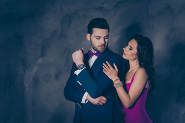 Portrait of harsh brutal handsome man with bristle in tuxedo with bowtie and charming pretty brunette woman with hairstyle attractive couple looking at each other isolated on grey background