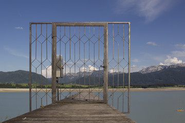 Looking through a locked lattice door onto a wooden boat dock across the dry Forgensee in the direction of the Alps