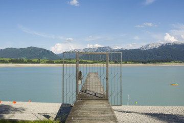 Looking through a locked lattice door onto a wooden boat dock across the dry Forgensee in the direction of the Alps