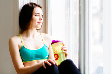 Fototapeta na wymiar Pretty athletic girl with short brown hair sitting near window with bottle and looking out.