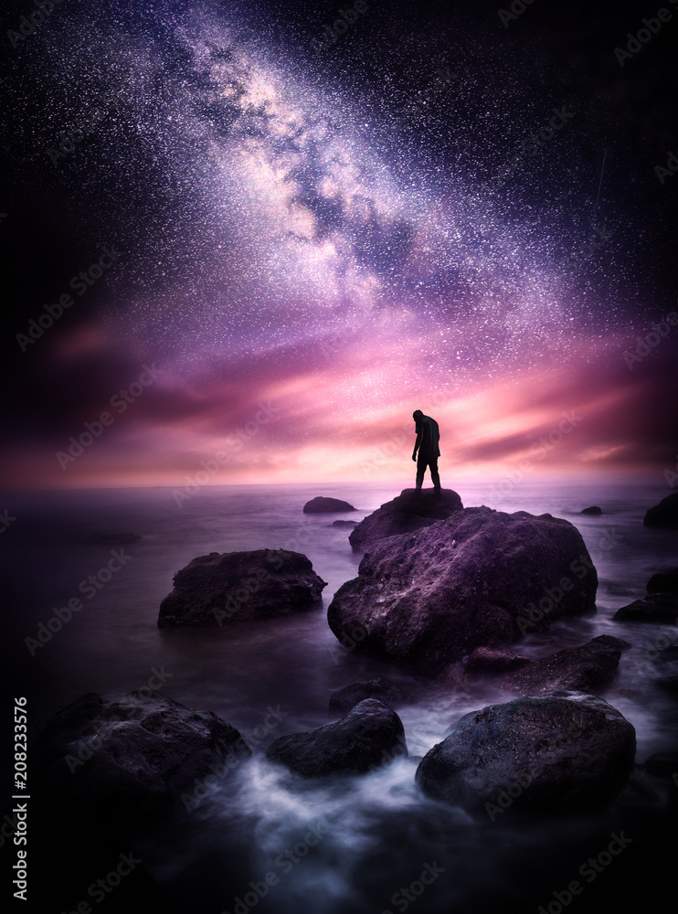 Wall mural night time sea landscape with the milky way. a man stands on a rocky shore line with the stars above