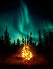 Fotobehang Noorderlicht Een warm en gezellig kampvuur in de wildernis met bosbomen als silhouetten op de achtergrond en de sterren en het noorderlicht (Aurora Borealis) die de nachtelijke hemel verlichten. Foto composiet.