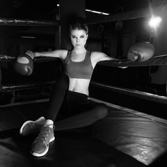 Full body portrait of boxer woman with long dark hair pulled back in pony tail, wearing pink sports bra, leggings, trainers and red boxing gloves, sitting in corner of ring and leaning on ropes.