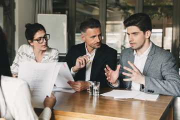 Group of business people in formal suits sitting at table in office, and talking with young woman about work during job interview - business, career and placement concept - obrazy, fototapety, plakaty
