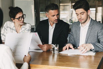 Group of caucasian people in formal suits sitting at table in office, and talking with young woman...