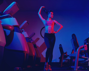 Waist up portrait of young attractive brunette woman dressed in sports clothes illuminated by faint pink light. Gorgeous confident slim female fitness model wearing top and leggings posing at gym.