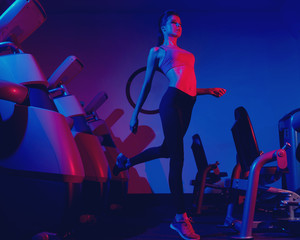 Young slim brunette woman dressed in sports apparel jumping among rows of exercise machines. Female model wearing leggings, top and trainers illuminated by pink and purple light posing at gym.