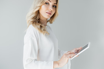 Fototapeta premium side view of young woman in white clothing with tablet in hands on grey backdrop