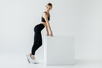side view of sportive woman in black sportswear standing near white cube on grey background