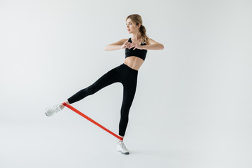 young athletic woman exercising with rubber tape on legs isolated on grey