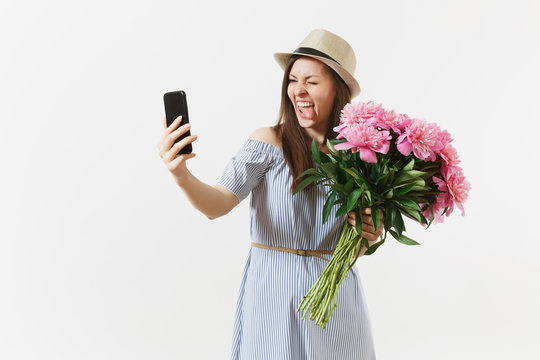 Young woman in blue dress, hat holding bouquet of beautiful pink peonies flowers, doing selfie on mobile phone isolated on white background. St. Valentine's, International Women's Day holiday concept.