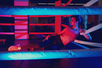 Seductive female fitness model with pony tail hairstyle, dressed in sports bra and leggings, illuminated by dim neon light, lying on left side of her body on boxing ring and leaning on ropes.