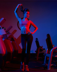 Waist up portrait of young attractive brunette woman dressed in sports clothes illuminated by faint pink light. Gorgeous confident slim female fitness model wearing top and leggings posing at gym.