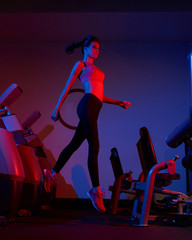 Young slim brunette woman dressed in sports apparel jumping among rows of exercise machines. Female model wearing leggings, top and trainers illuminated by pink and purple light posing at gym.