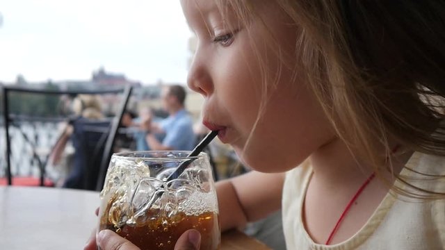 Funny little child girl portrait enjoy making bubbles in glass of juice with straw at street restaurant table
