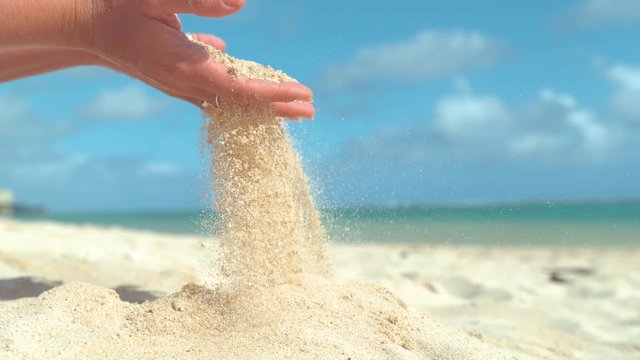 SLOW MOTION, CLOSE UP, COPY SPACE: Gentle summer wind blows tiny grains of white sand out of playful woman's soft hand. Unrecognizable female tourist plays with hot sand on the exotic sunny beach.