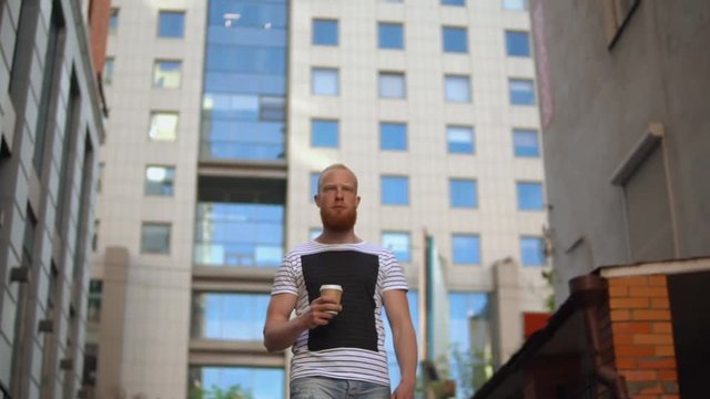 handsome guy going to the work slow motion successful man holding to go coffee. on the background modern building. unban city view in summer season