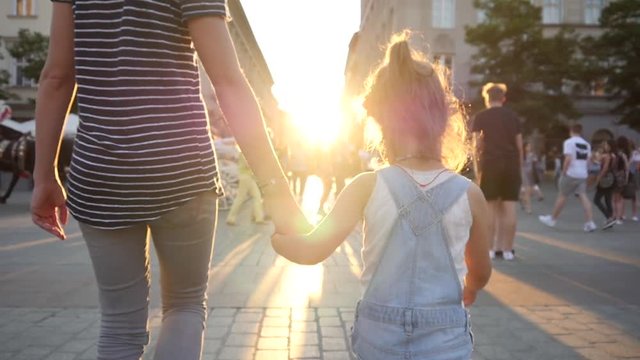 Mother walk with child girl daughter holding her hand in hand in a city in sun rays
