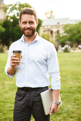 Portrait of successful entrepreneur man in white shirt, standing with takeaway coffee and silver laptop during walk in park