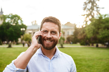 Happy photo of smiling adult man 40s in white shirt, looking at you while talking on black smartphone during walk in green park