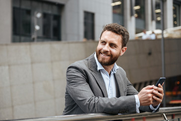 Photo closeup of adult unshaved businesslike man in gray suit looking aside and holding cell phone, while standing in front of modern office building