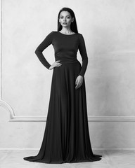 Naklejka premium Young woman with long brunette hair dressed in elegant evening teal maxi dress with long sleeves posing in studio. Gorgeous female model standing against white wall decorated with classic moldings.