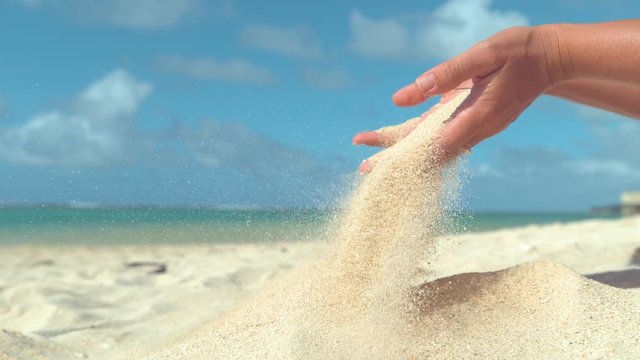 SLOW MOTION, CLOSE UP, COPY SPACE: Summer breeze sweeps away white sand out of playful woman's gentle hands. Beautiful shot of particles of sand falling out of girl's grasp on a sunny exotic beach.