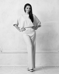 Young brunette woman with long straight hair wearing trendy short sleeved blue jumpsuit and nude heeled shoes posing against white wall on background. Female model demonstrating fashionable outfit.