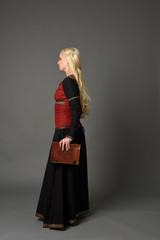 Fototapeta premium full length portrait of pretty blonde lady wearing a red and black fantasy medieval gown, holding a book. standing pose on grey background.