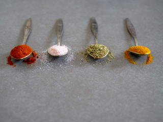 Various dry herbs and spices in vintage silver teaspoons. Flat lay of basil, turmeric, paprika and Himalayan pink salt on gray background.