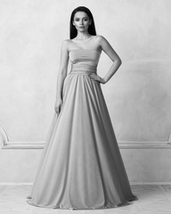 Fototapeta premium Beautiful long haired young woman dressed in stylish turquoise blue bandeau maxi dress posing against white wall on background. Elegant brunette female model demonstrating evening outfit in studio.
