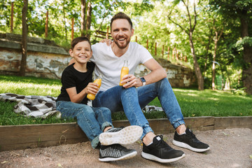 Happy young father sitting with his little son outdoors drinking juice.