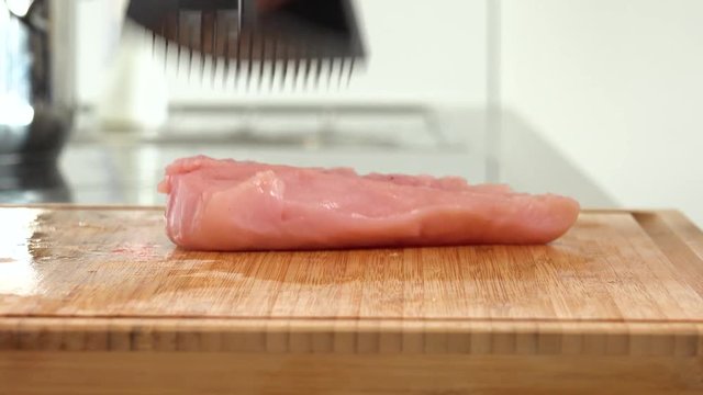 A woman pounds a chicken breast on a chopping board in a kitchen - closeup