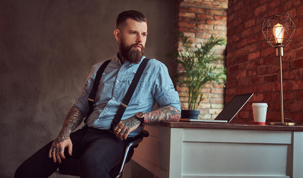 Handsome tattooed hipster in a shirt and suspenders sitting at the desk with a computer, looking out the window in an office with loft interior.