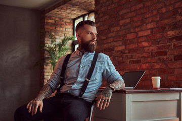 Handsome tattooed hipster in a shirt and suspenders sitting at the desk with a computer, looking...