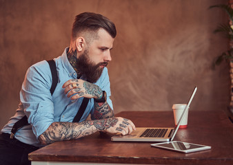 Handsome tattooed hipster in a shirt and suspenders sitting at the desk, working on a laptop, in an...
