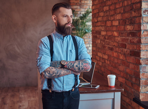 Old-fashioned tattooed hipster in a shirt and suspenders, standing near a desk with a laptop, looking out the window in an office with loft interior.