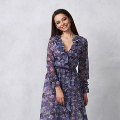 Full body portrait of attractive young woman posing in elegant maxi chiffon evening dress with motley pattern, long sleeves and frills. Brunette girl standing against white wall on background.