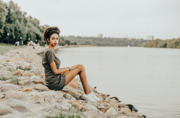 Fototapeta na wymiar Charming African-American girl is sitting on a stony quay of a river with the park and a walkway behind her; young curly Brazilian female is sitting on a stone embankment on a summer evening