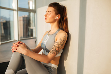 Portrait of young lady in sporty top and leggings sitting on yoga mat and listening music in earphones while dreamily closing her eyes at home with big beautiful windows isolated