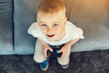 Fototapeta premium Bright buttons. New modern games console in the hands of a pretty little boy sitting on a sofa