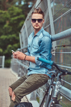 Handsome hipster with stylish haircut in sunglasses resting after riding on a bicycle, holds a smartphone.