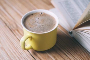 Yellow mug with hot black coffee on a wooden table with an old book.