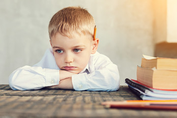 Naklejka premium Bored pupil. Tired bored little boy sitting sadly at the table with books by his side